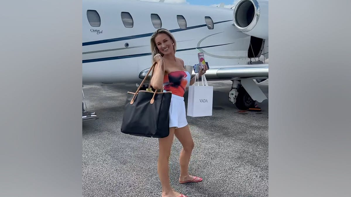 Brittany Aldean walks on an airport tarmac carrying bags and a drink while a private jet sits behind her.