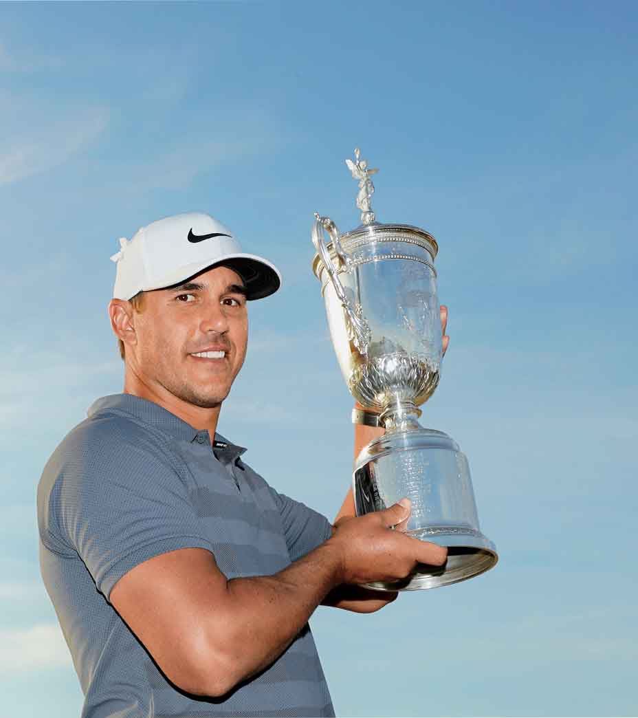 A male golfer in a gray shirt and white cap smiles while holding up a large silver trophy, celebrating among the winners at the U.S. Open under a clear blue sky.