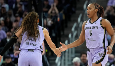 Two female basketball players in purple Golden State uniforms are giving each other a high five mid-game in an arena filled with spectators.