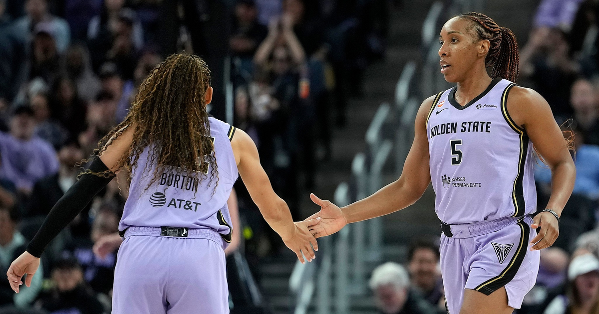Two female basketball players in purple Golden State uniforms are giving each other a high five mid-game in an arena filled with spectators.