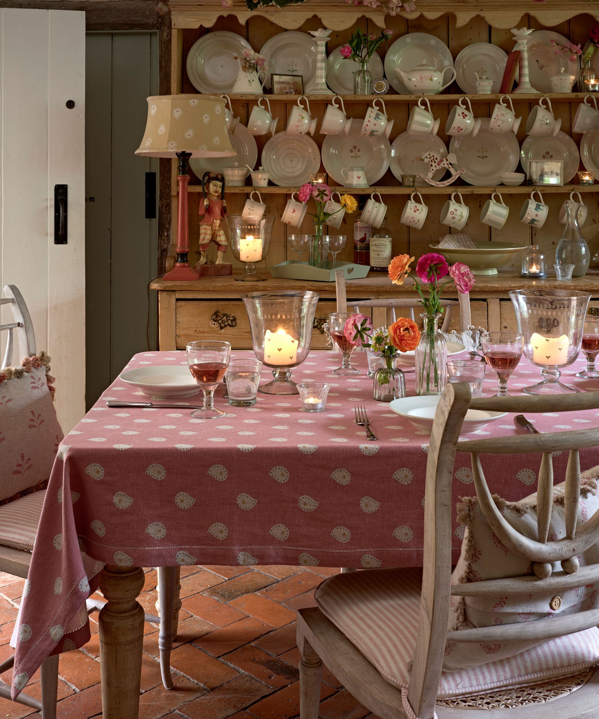 Dining table with a pink patterned tablecloth and hurricane candles, set against a wooden Welsh dresser stacked with cream plates and hanging teacups
