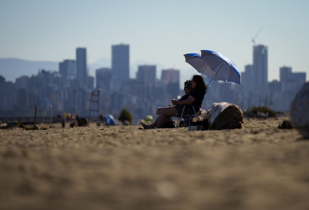 People sit under umbrellas at Locarno Beach in Vancouver. Skyscrapers stand tall in the background.