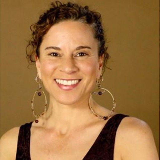 headshot of woman smiling at the camera with hair tied back wearing large hooped earrings and a black vest