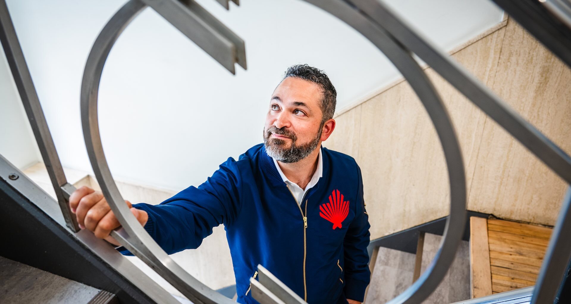 A photo of Daniel Sax shot through a circular piece of ironwork on a stairway balustrade. He's looking off-camera, and is wearing a dark blue jacket bearing his company's logo.