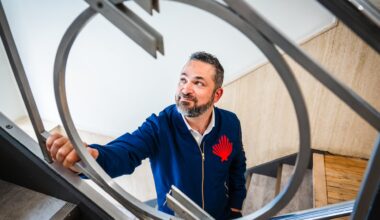 A photo of Daniel Sax shot through a circular piece of ironwork on a stairway balustrade. He's looking off-camera, and is wearing a dark blue jacket bearing his company's logo.