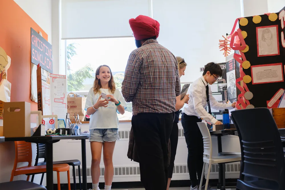 A young girl speaks with a man in a red turban with heritage fair projects in the background
