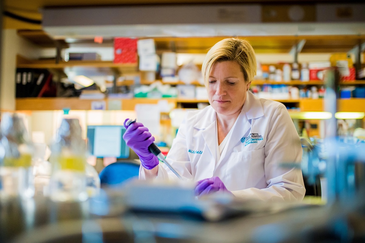 A woman with light skin tone and blond hair wears a white lab coat and purple latex gloves. She is focused on a lab task.