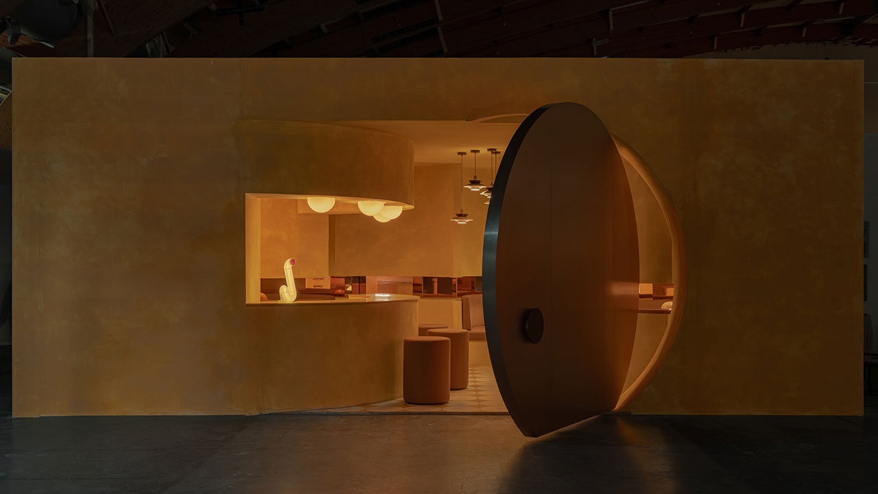 A modern interior with warm yellow lighting features a large circular door, a counter with stools, pendant lights, and a decorative duck statue on the counter.