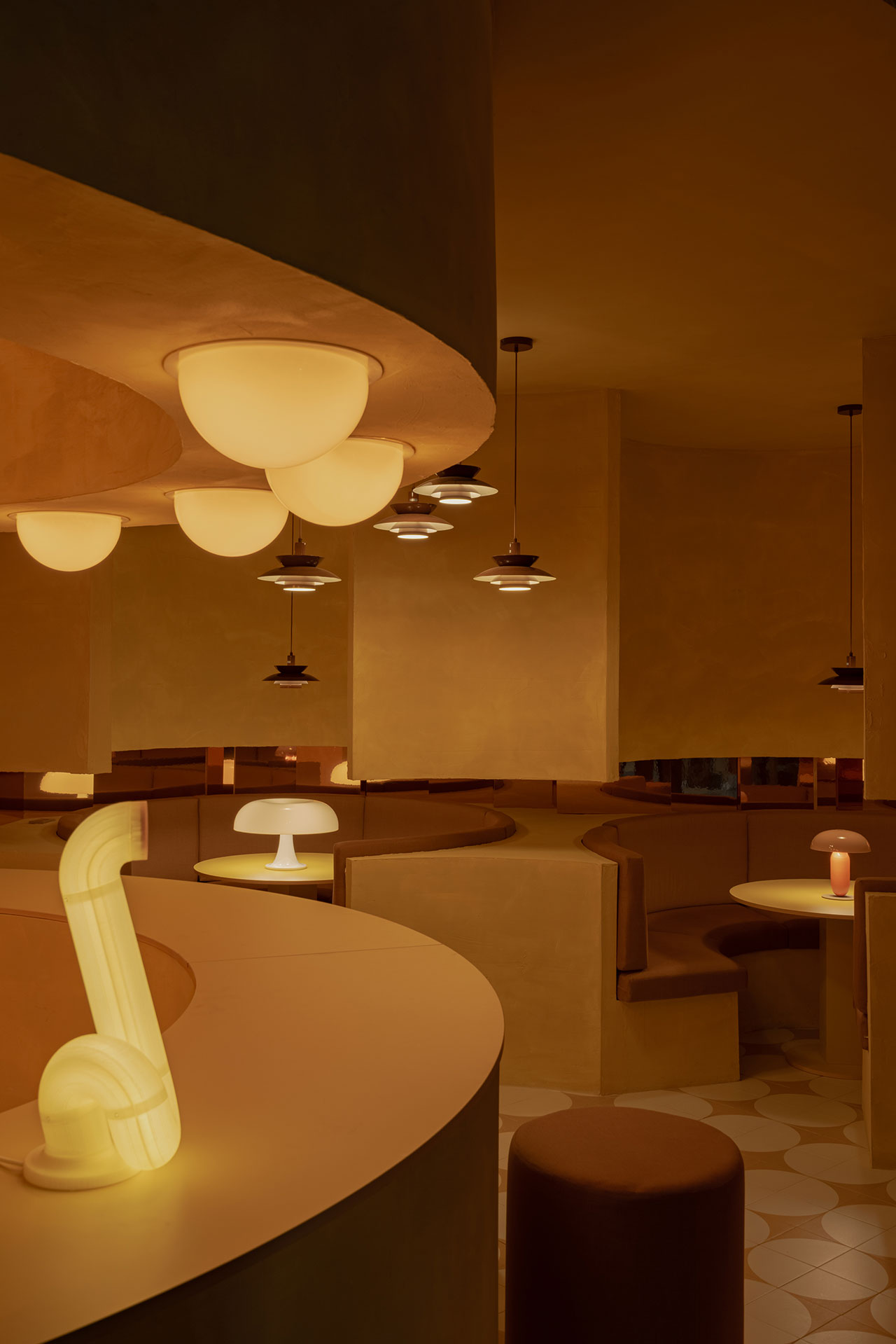 A dimly lit restaurant interior with curved booths, round tables, and modern amber and white lamps creating a warm, golden atmosphere.