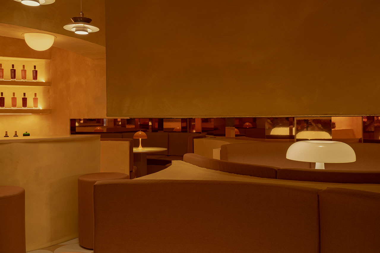 A modern, dimly lit restaurant interior with curved brown seating, round tables, and minimal decor, featuring shelves with bottles and globe-shaped table lamps.