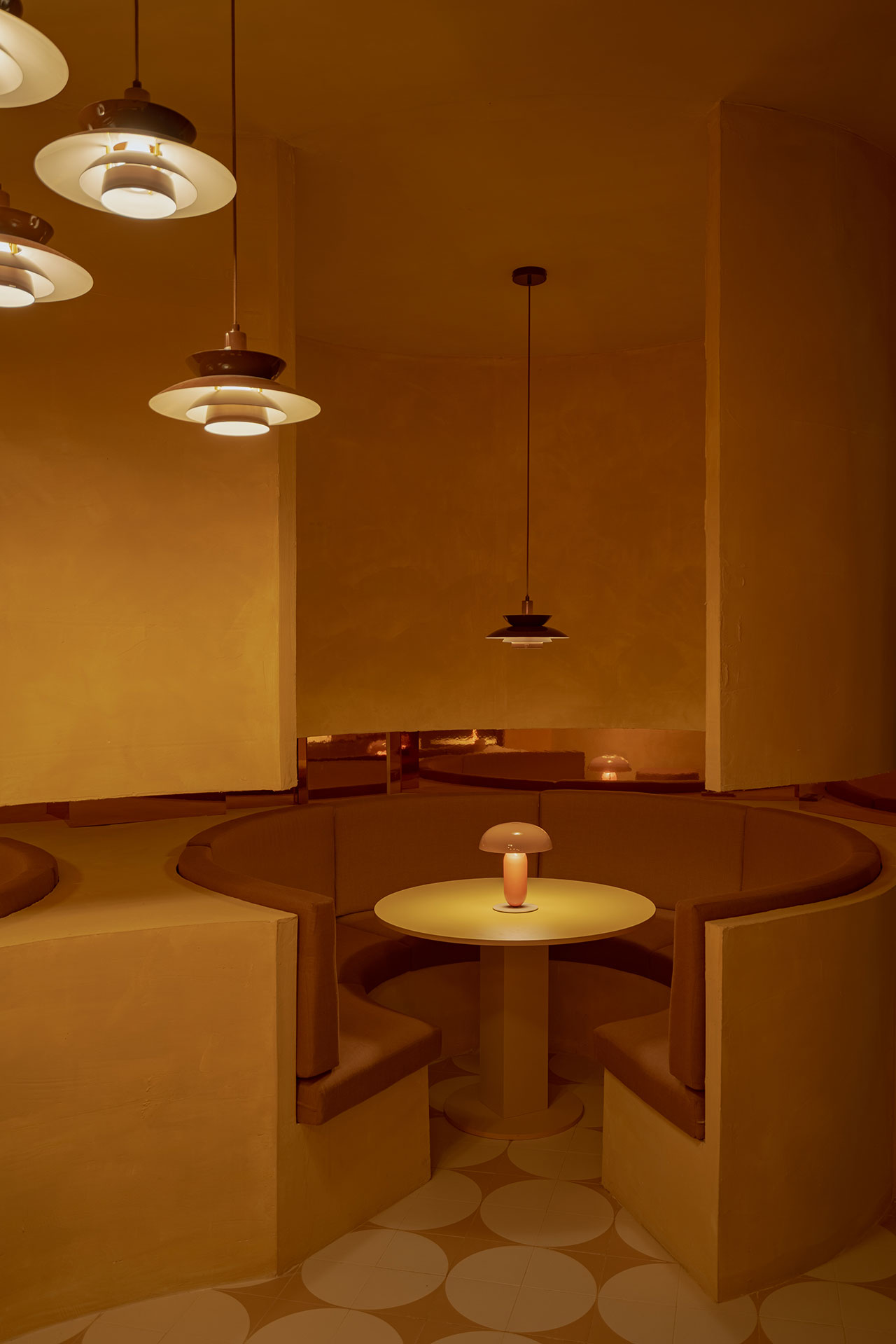 A round booth with brown cushioned seats surrounds a circular table with a small lamp, set in a warmly lit, yellow-toned interior beneath several hanging lights.