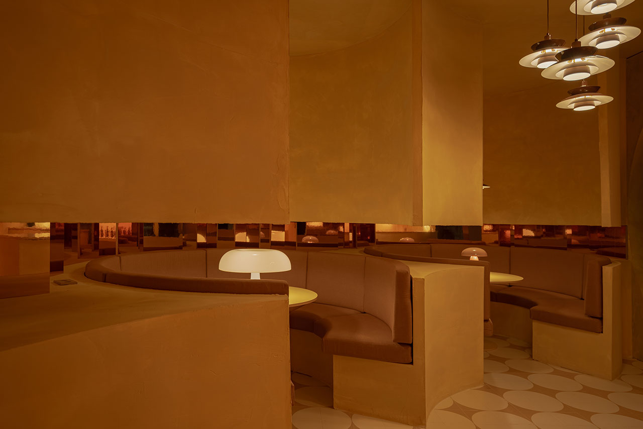 Curved booth seating and round tables in a restaurant with warm brown tones, minimalist decor, and pendant lights hanging from the ceiling.