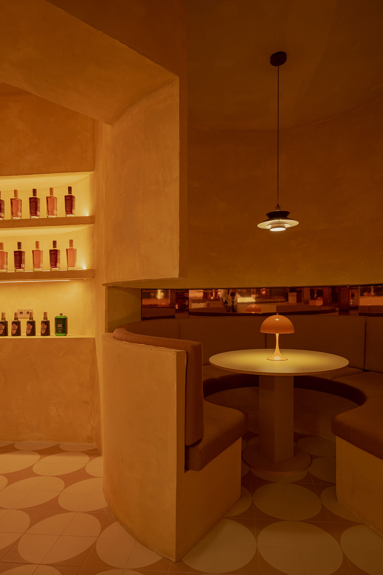 A dimly lit, modern restaurant booth with a round table, curved seating, a small lamp, and shelves of liquor bottles against a warm beige interior.
