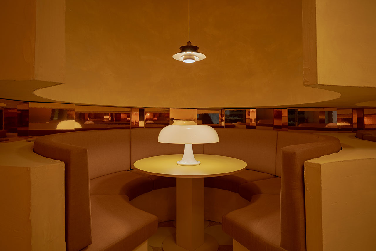 A round booth with brown cushioned seating surrounds a circular table with a white lamp, under a ceiling light, in a room with warm yellow and brown tones.