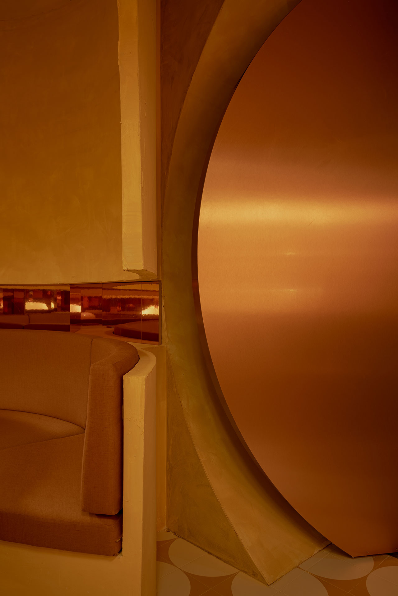 A round metallic door with a warm bronze finish next to a curved beige sofa and a patterned floor in a modern interior space.
