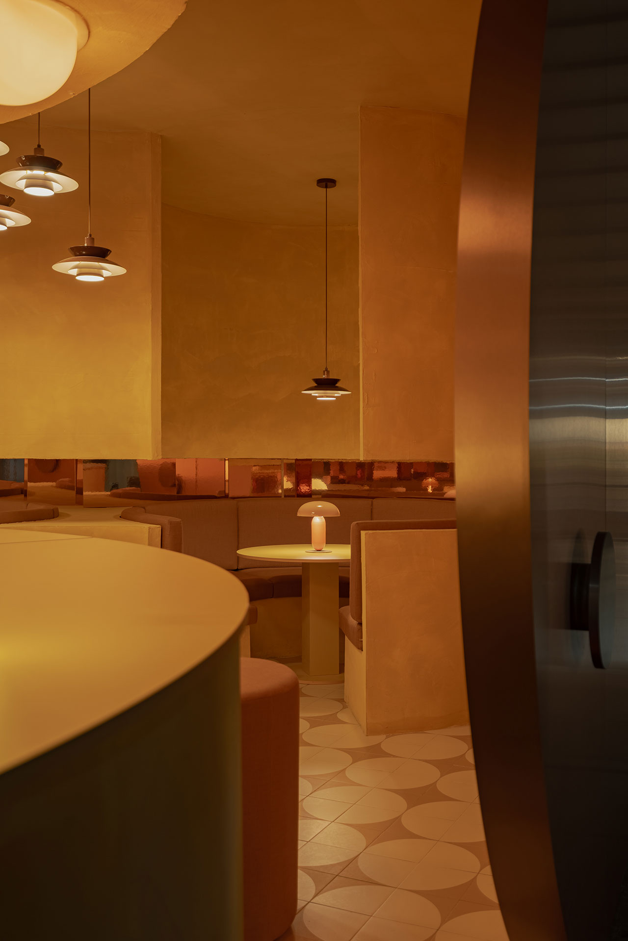 A warmly lit, modern restaurant interior with curved booths, pendant lights, and geometric tiled flooring in shades of beige and gold.