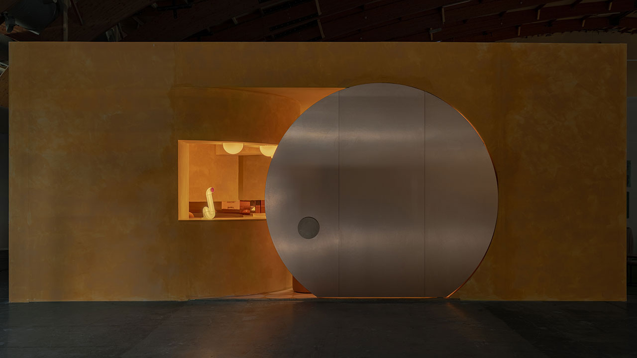 A minimalist interior features a large circular metallic sliding door next to a rectangular window opening, set in a textured yellow wall.