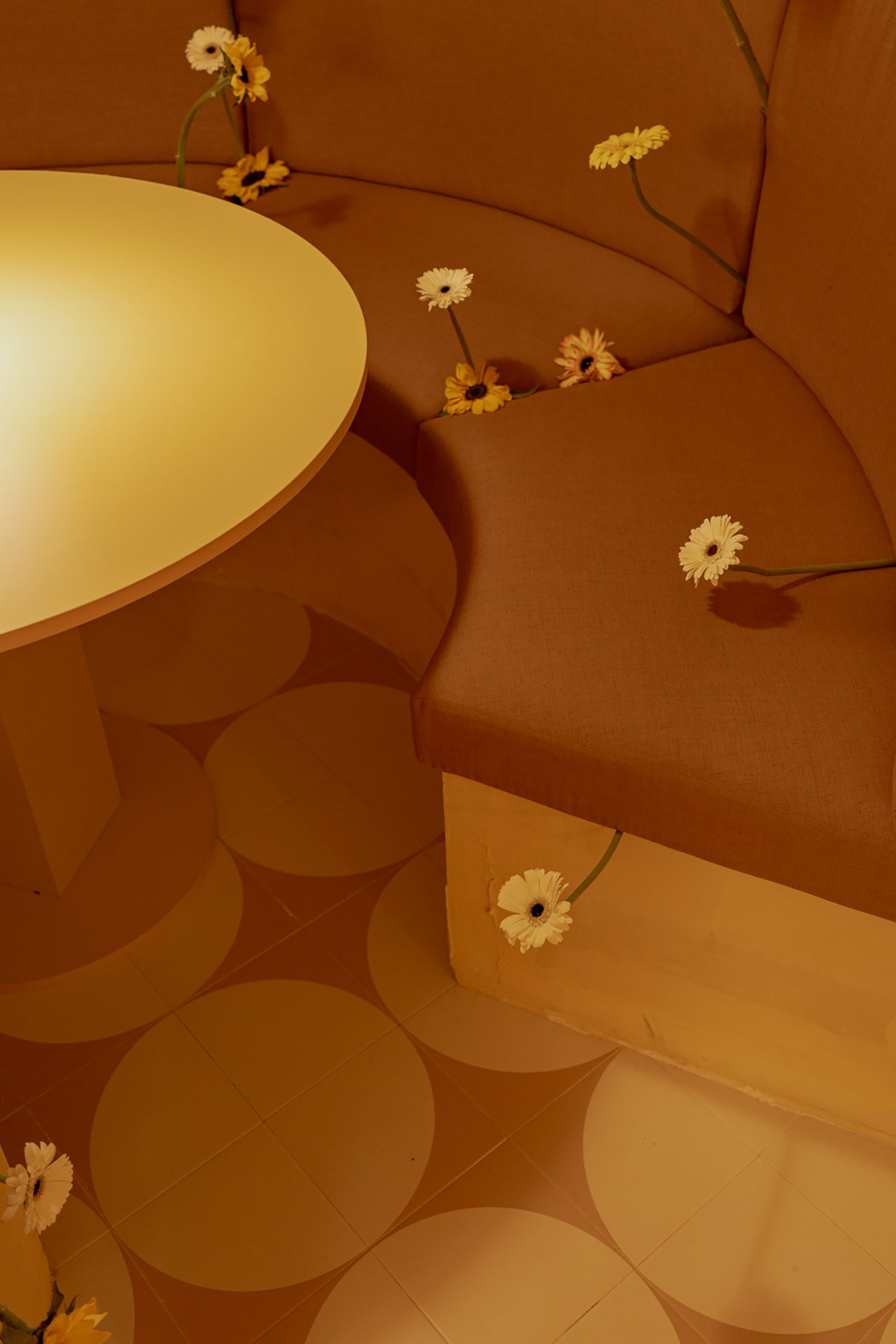 A curved brown bench and round table with white and yellow flowers inserted between the cushions, set on a tiled floor with a circular pattern.