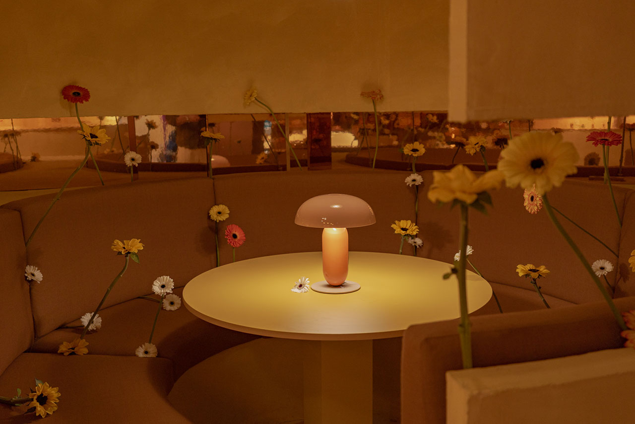 A round table with a mushroom-shaped lamp sits in a curved booth surrounded by yellow and white daisies in a softly lit room.