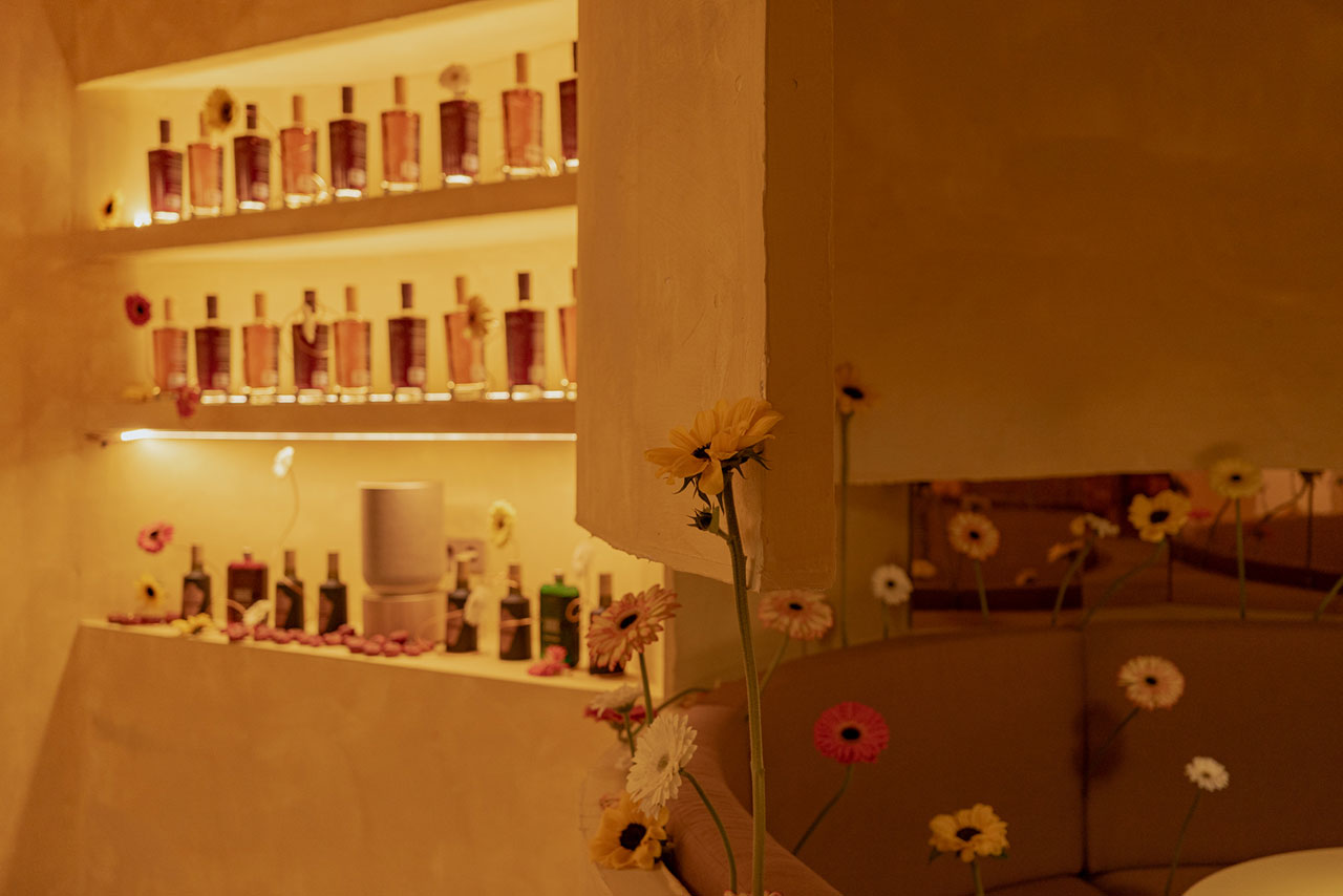 Shelves display bottles and a speaker against a yellow wall, with single-stem flowers placed around a curved seating area.