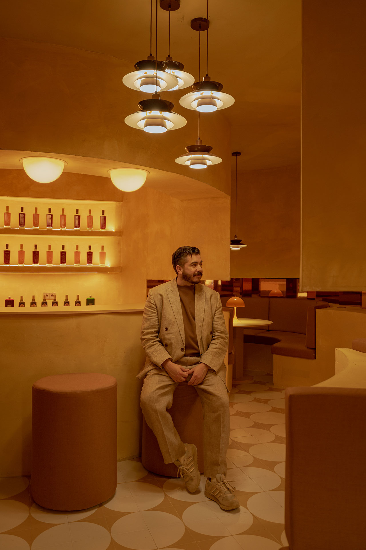 A man in a beige suit sits on a stool in a warmly lit, modern room with round lights, tiled floor, and shelves displaying bottles.