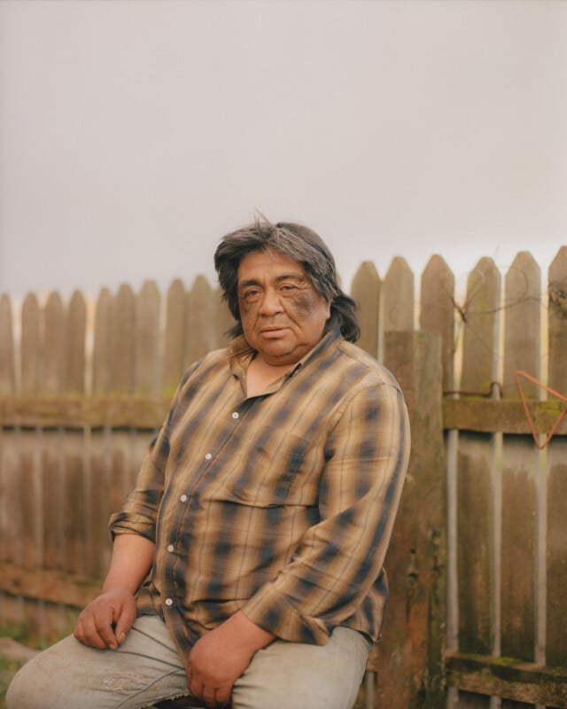 A man with long dark hair wearing a brown plaid shirt and light jeans sits in front of a wooden fence outdoors, looking directly at the camera with a neutral expression.