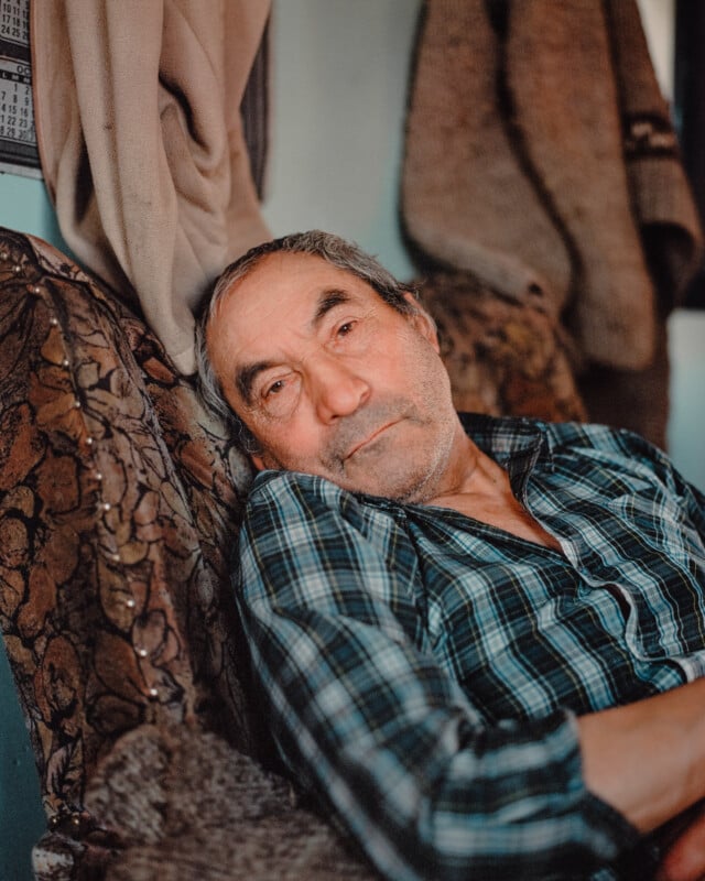 An elderly man with gray hair and a beard, wearing a blue plaid shirt, reclines on a patterned chair indoors, looking thoughtfully at the camera. Coats or blankets hang in the background.