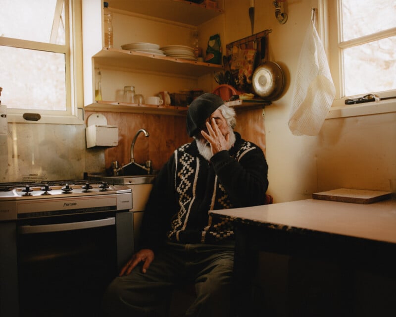An elderly person with a white beard sits in a small, warmly lit kitchen, wearing a patterned sweater and cap, with one hand covering their face. Shelves, dishes, and a stove are visible in the background.