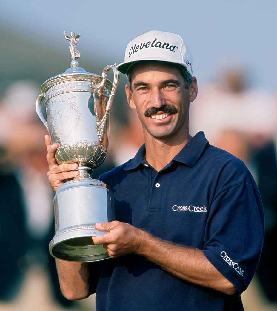 A smiling man in a blue polo shirt and white cap holds a large silver trophy with both hands, celebrating among the winners at Shinnecock Hills, with a blurred crowd in the background.