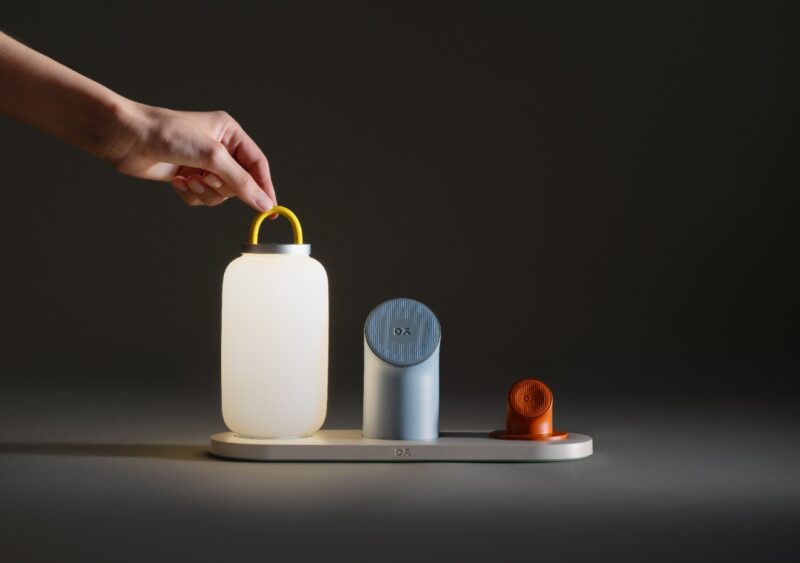 A hand lifts a lantern-shaped light with a yellow handle from a minimalist DailyObjects docking station holding two other small objects on a neutral background.