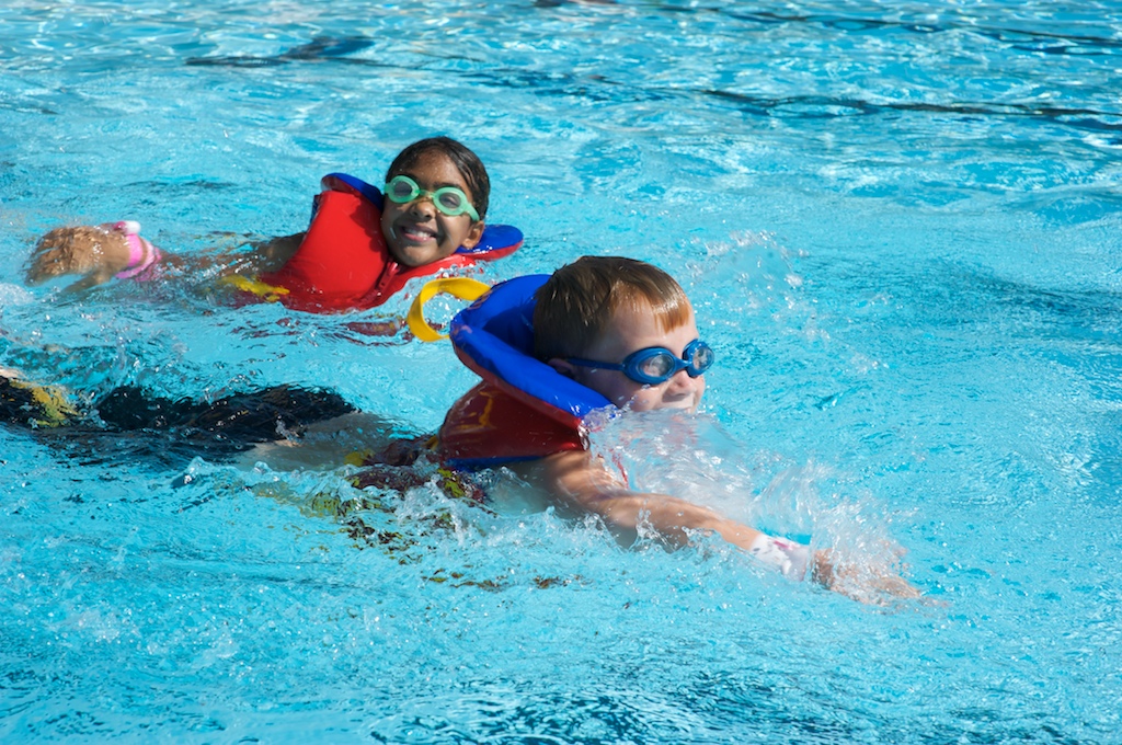 Two children swimming in a pool while wearing life jackets.