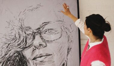 A woman with dark hair tied back, wearing a pink vest over a white shirt, reaches up to touch a large, expressive sketch of a face on a canvas.
