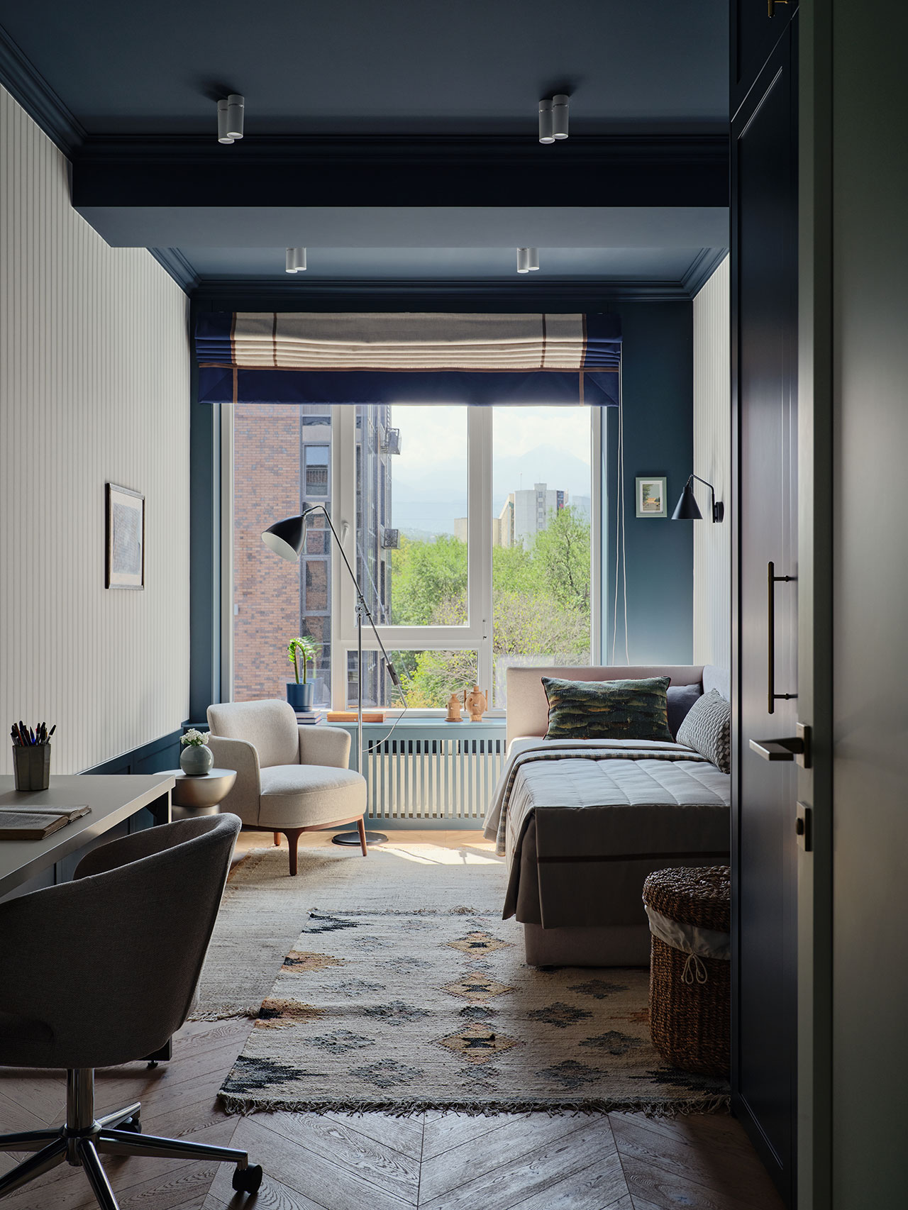 A modern, neatly arranged bedroom with a single bed, armchair, desk, large window, and city view. Natural light brightens the space, decorated in neutral and blue tones.