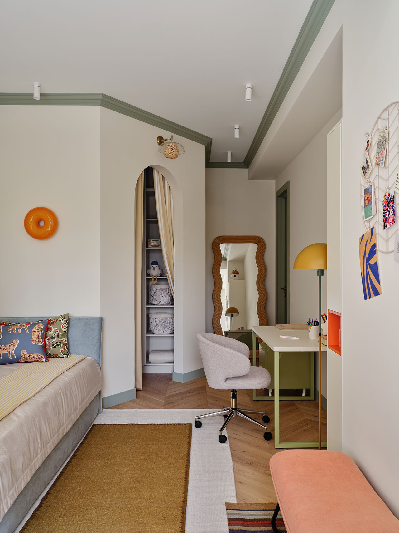 A modern bedroom features a daybed, a desk with a chair and lamp, a large mirror, an orange bench, and an open curtained closet with shelves. The decor includes warm and pastel tones.