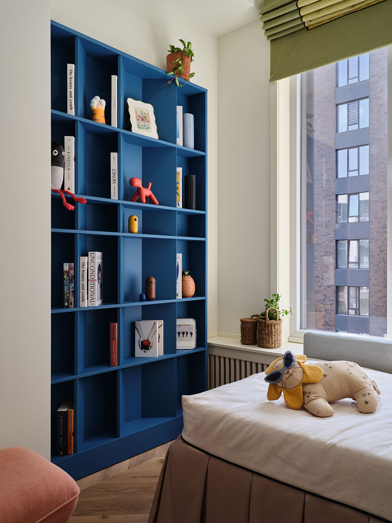 A modern bedroom with a blue bookshelf filled with books and decorative objects, a bed with a plush lion toy, and a large window showing a building outside.