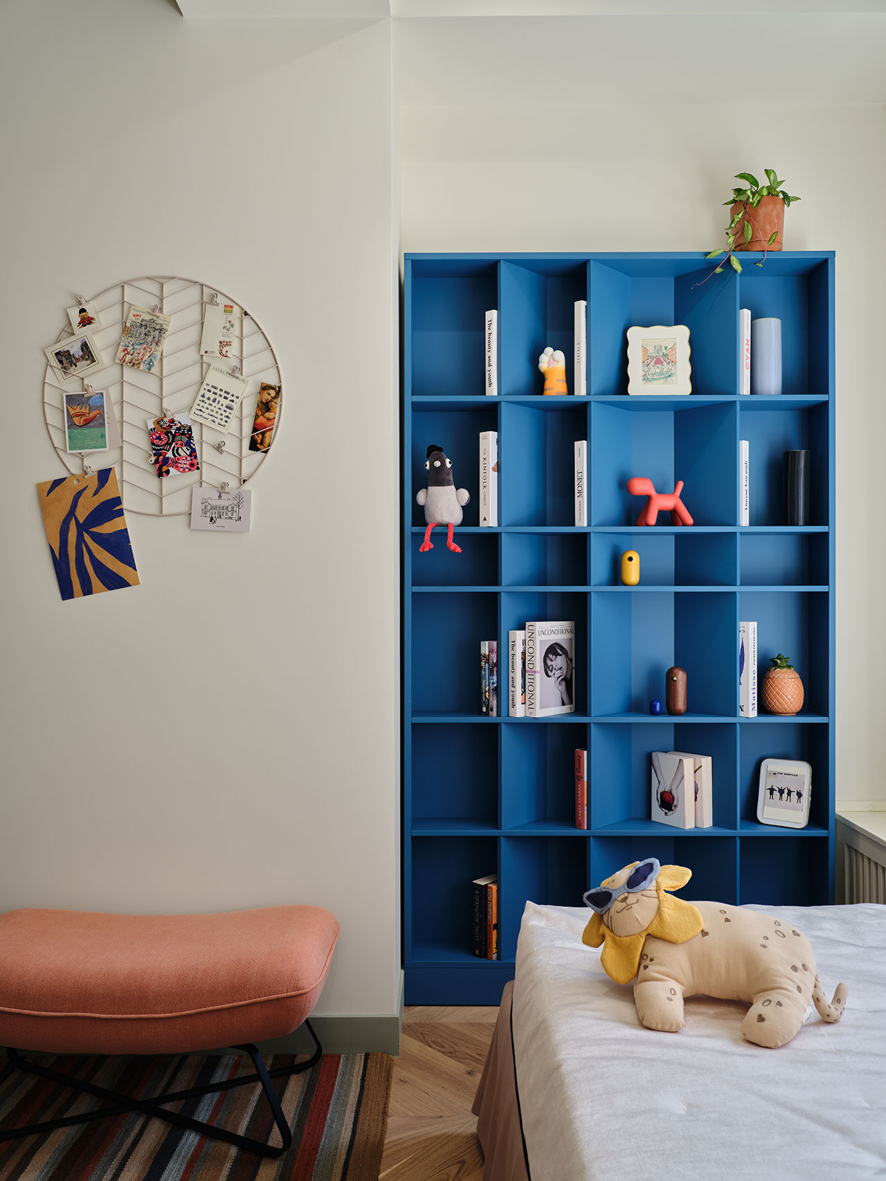 A modern room with a blue bookshelf holding books and decor, a circular wall organizer with photos, a coral bench, a striped rug, and a stuffed animal on a bed.