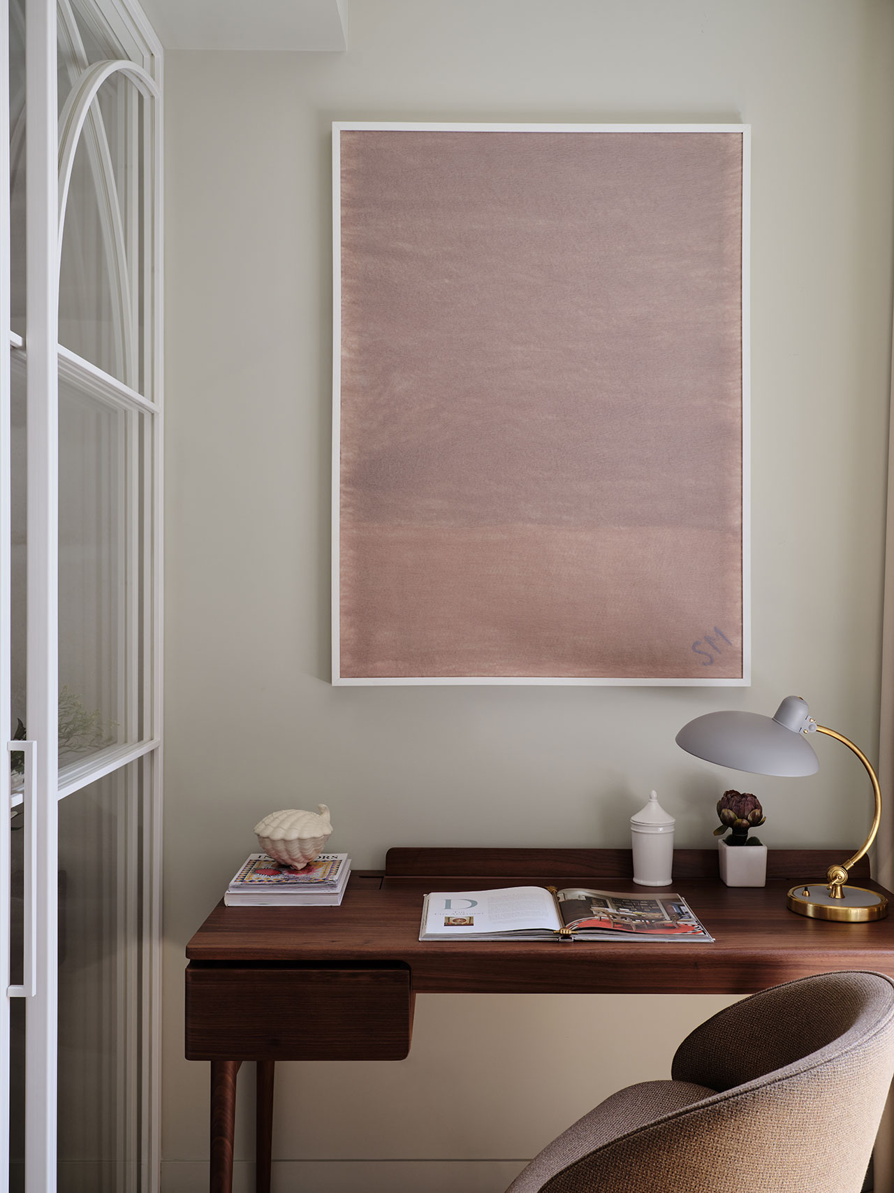 A small wooden desk with an open book, lamp, decorative items, and a chair, set against a light wall with a large framed minimalist artwork above.
