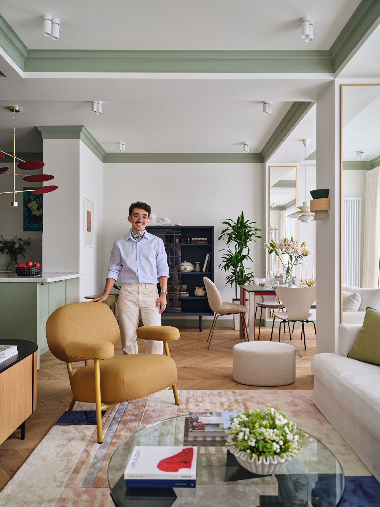 A man stands in a modern, stylish living room with light-colored walls, green accents, contemporary furniture, and decorative plants.