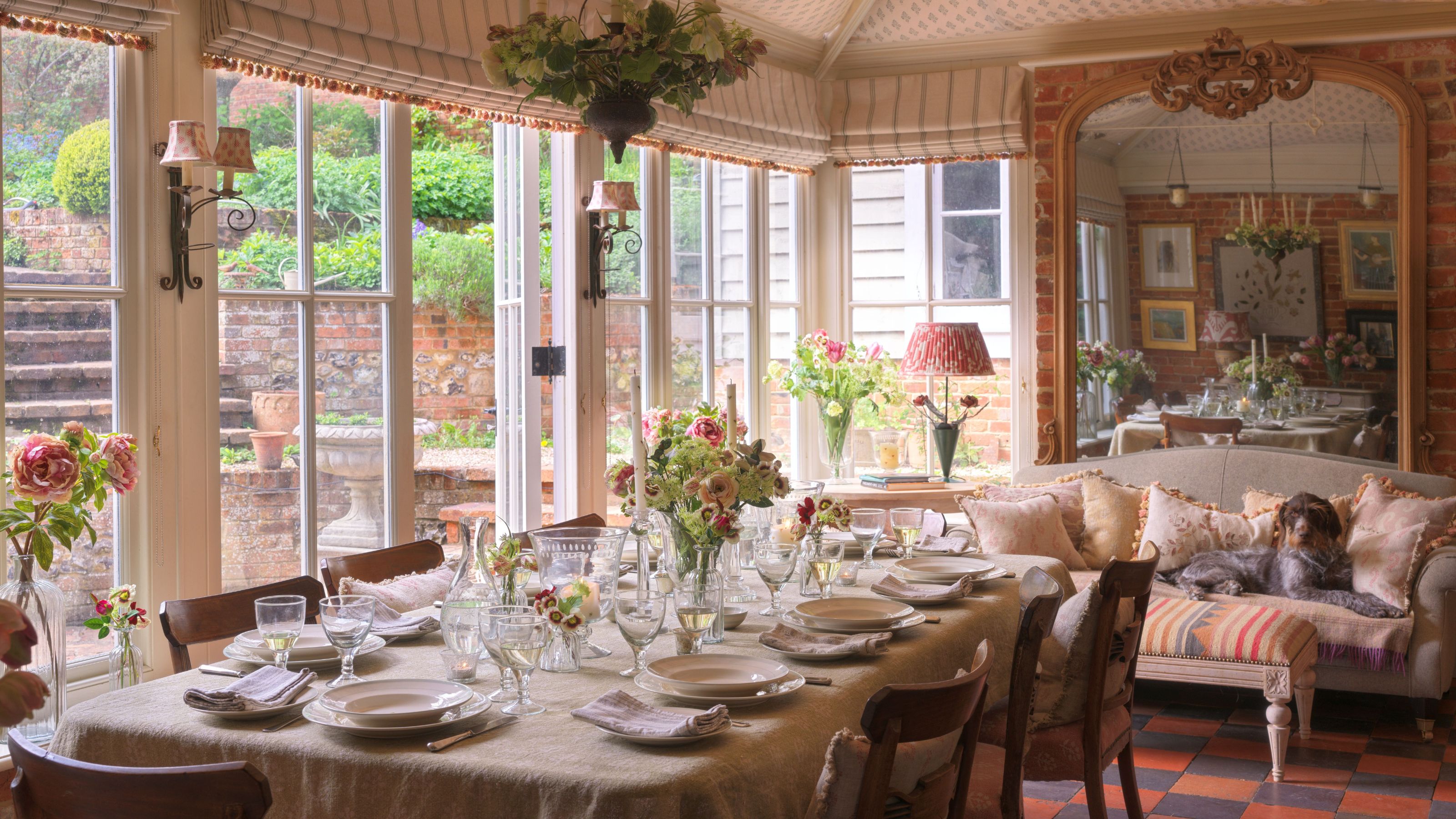 Sunlit country dining room with an elegant table setting, floral centerpieces, and a dog resting on a cozy sofa in the background.