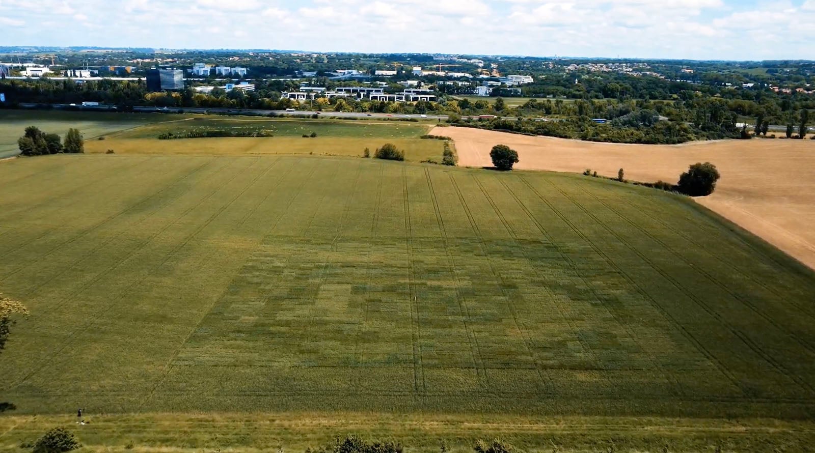 A large green field with faint rectangular patterns visible in the grass, seen from above. Other fields, trees, and a distant town with buildings are in the background under a blue sky with clouds.