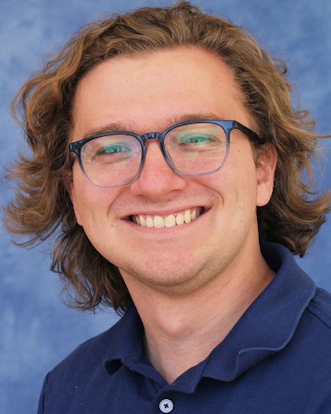 Smiling young man with longish, tousled light brown hair wearing eyeglasses.