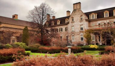 A large historic stone building with tall windows and dormers stands behind landscaped gardens with brown shrubs, winding paths, and a sundial under cloudy skies.