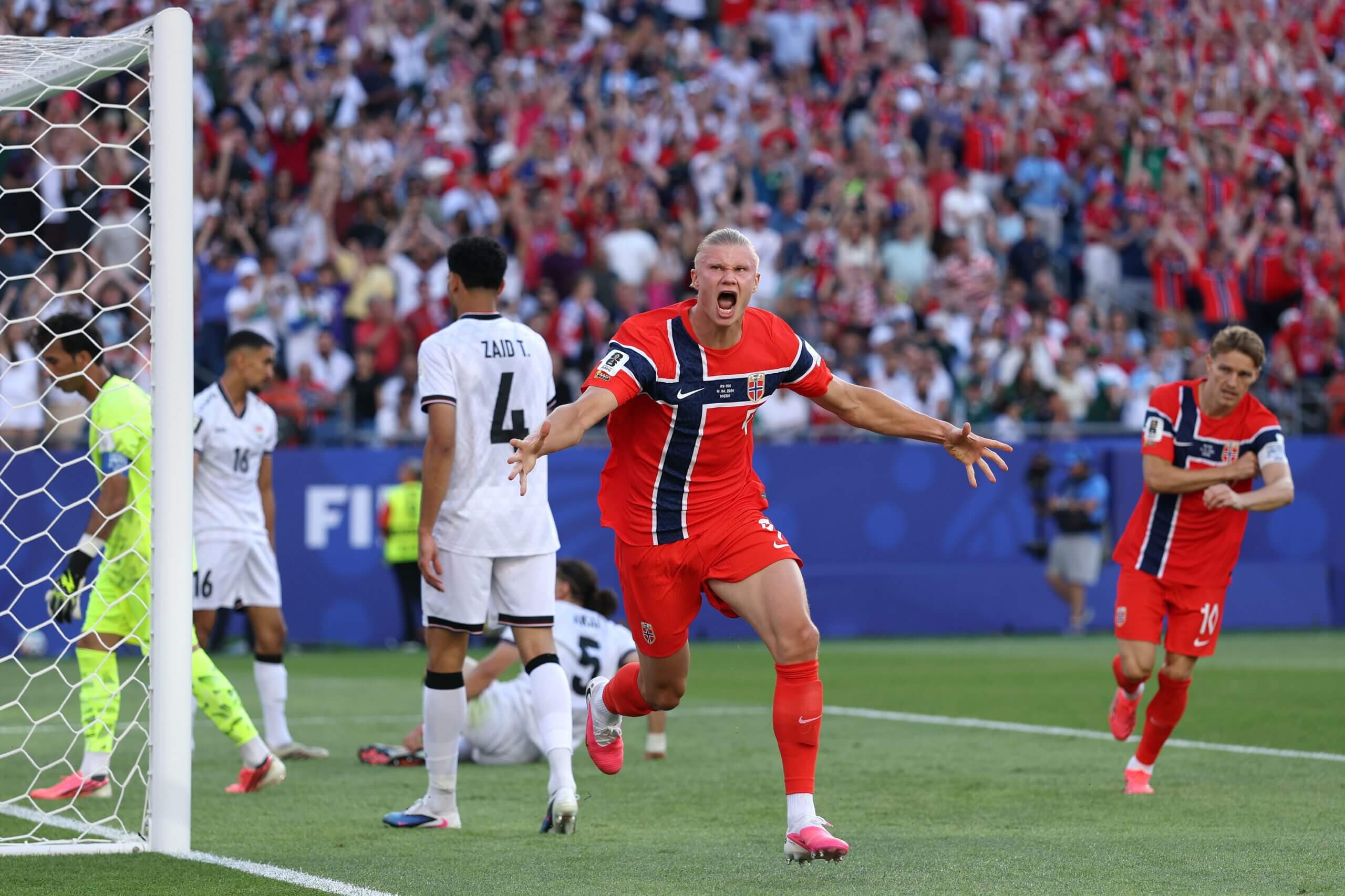 Erling Haaland celebrates scoring Norway's first goal during the World Cup against Iraq and Norway at Gillette Stadium. He is running, arms outstretched, towards supporters as Iraq's goalkeeper and defenders slump around the six yard box