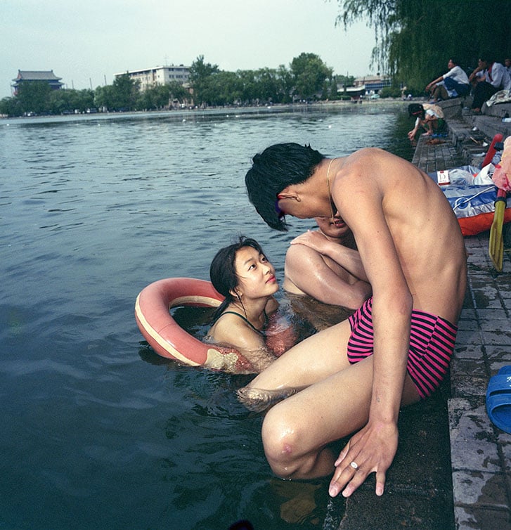 Swimming woman looks up at man sitting on the pool’s edge.