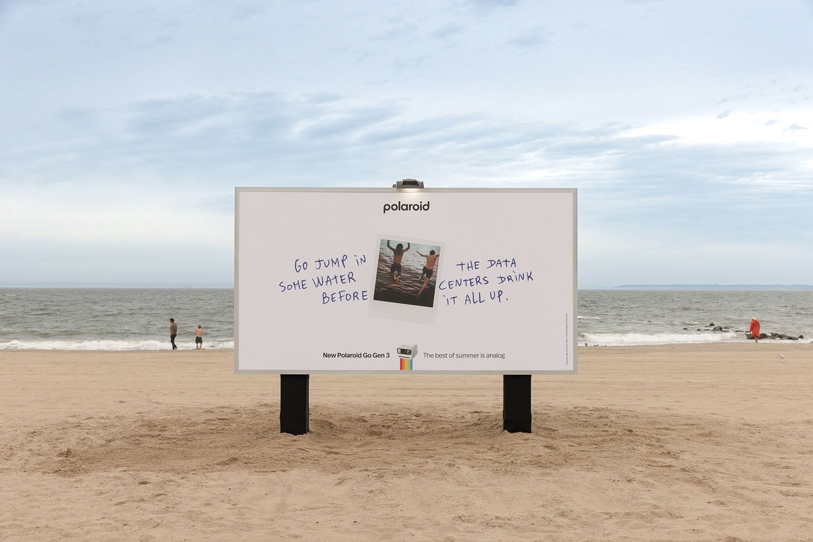 A Polaroid billboard on a sandy beach shows two instant photos above the text: "Go jump in some water before the data centers drink it all up." The ocean and a few people are visible in the background.