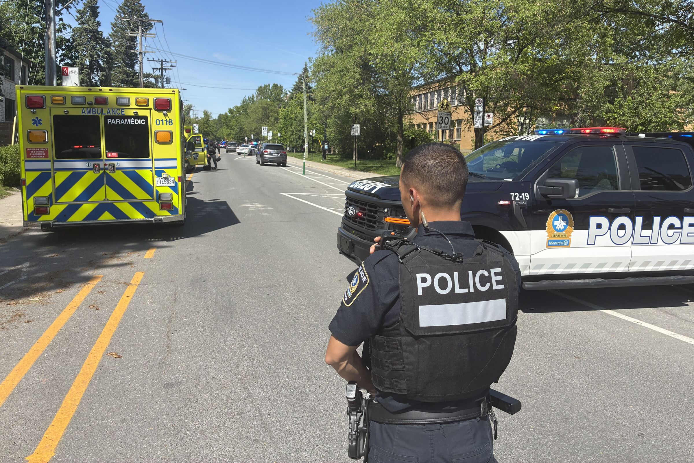 A police officer stands in a street near a police car and ambulance