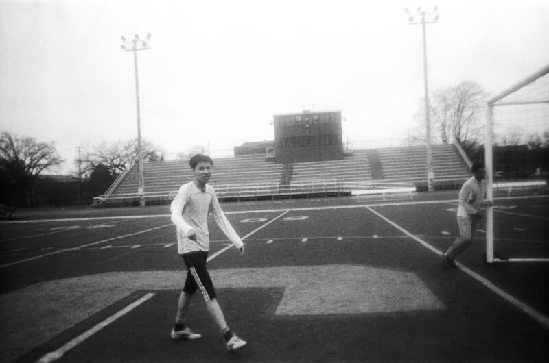 Two people play soccer on a field; one walks near the goalpost while the other stands in the goal. Bleachers and lights are visible in the background on an overcast day. The image is in black and white.
