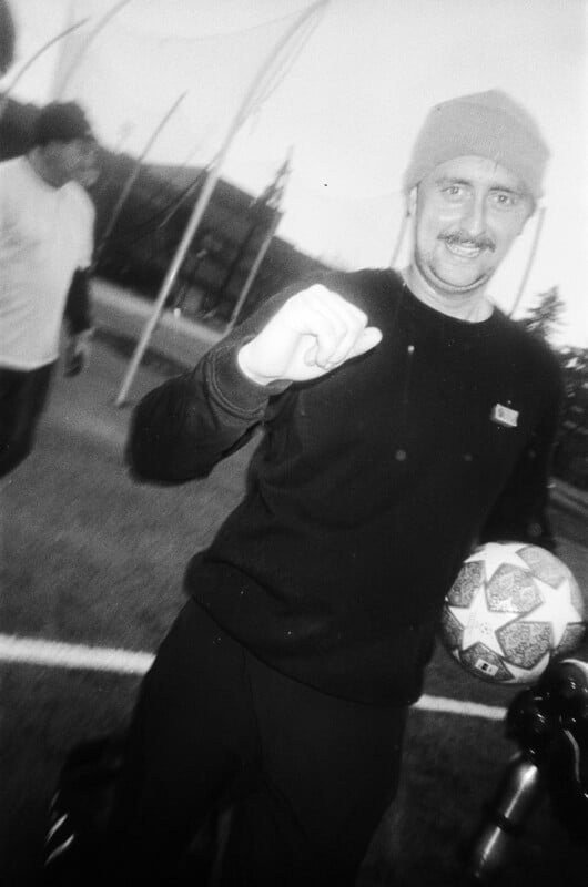 A man wearing a beanie and sweater smiles and raises his fist while holding a soccer ball on a field. Another person stands in the background. The image is black and white and slightly blurry.