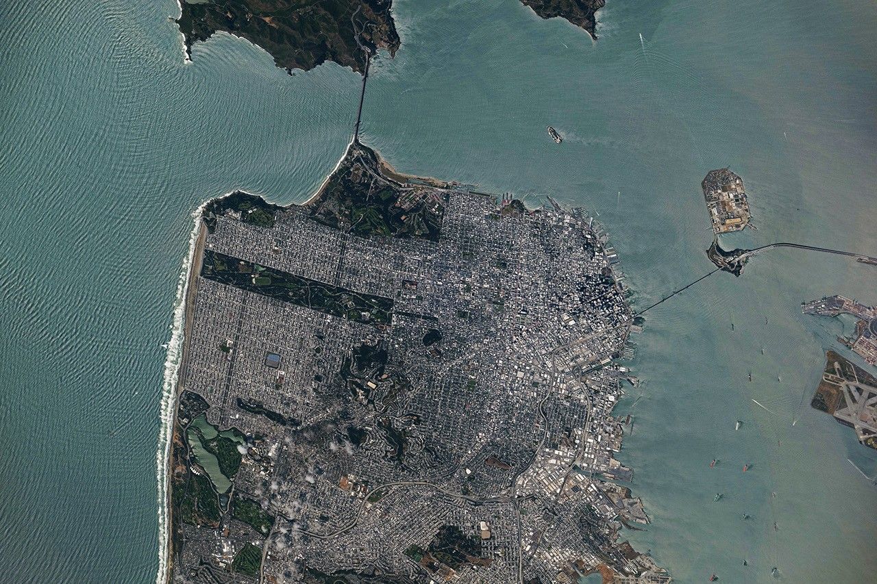 A top-down photo of San Francisco shows dense gray urban infrastructure interspersed with green parks. Waves and ships are visible in the surrounding blue-green water.