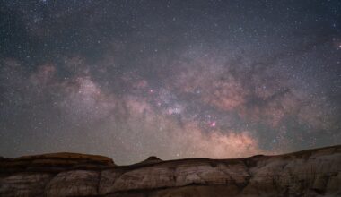 A star-filled night sky with the Milky Way galaxy glowing above rugged, rocky cliffs in the foreground. The scene highlights bright stars, cosmic dust, and soft, colorful nebulae stretching across the horizon.